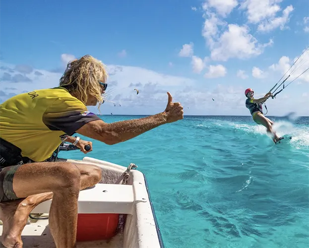 Persoon die leert kitesurfen op het water van Bonaire met een instructeur die haar begeleidt en een duim omhoog geeft. Begin met kitesurflessen of huur materiaal bij onze partner en geniet van speciale kortingen op pick-up verhuur bij Pickup Huren Bonaire.